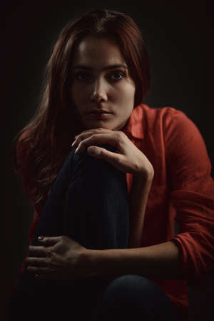 Young beautiful beautiful woman in coral shirt sitting, hugging her leg and looking pensively in camera on dark background, low key studio shotの写真素材