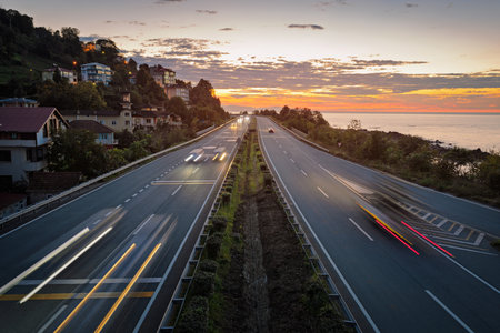 Highway along the sea coast at dusk. Light trails from vehicles. Houses on a hill.