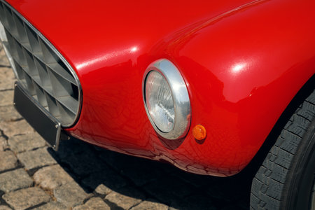 Close-up of a vintage red sports car's front fender, showing the chrome headlight and patterned grill on a cobbled street.の写真素材