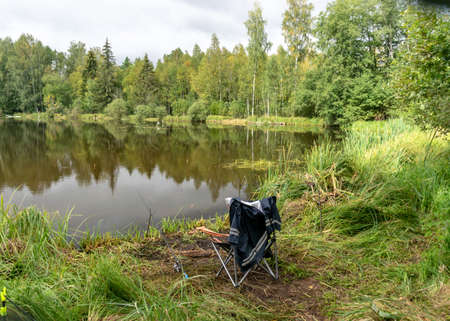 landscape with lake shore and fisherman's chair, calm lake water surface, tree reflections, cloudy day, fishing conceptの写真素材