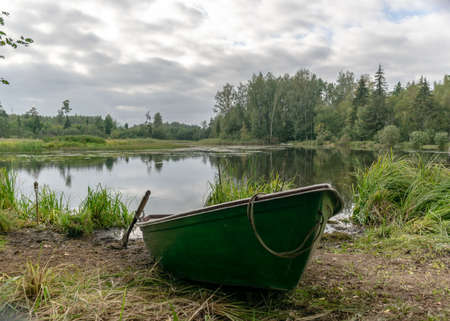 landscape with lake shore and boat, calm lake water surface, tree reflections, cloudy day, fishing conceptの写真素材