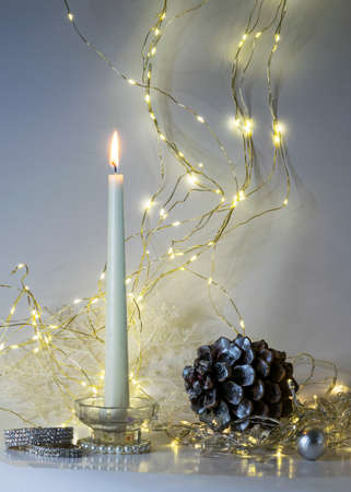 white burning candle, large cone and various Christmas decorations, illuminated background of a chain of white lights, Christmas waiting time, winter timeの写真素材