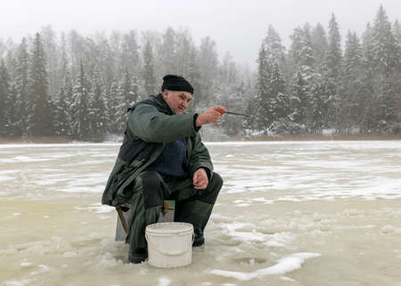 landscape with a man in winter who sits on the ice next to a hole and holds a small fishing rod. A fisherman is sitting on a frozen lake and fishing, winter dayの写真素材