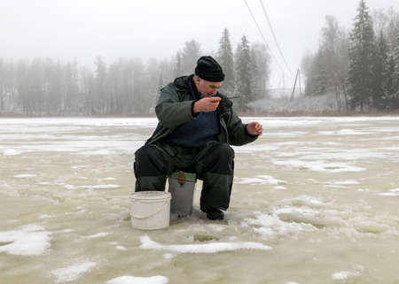 landscape with a man in winter who sits on the ice next to a hole and holds a small fishing rod. A fisherman is sitting on a frozen lake and fishing, winter dayの写真素材