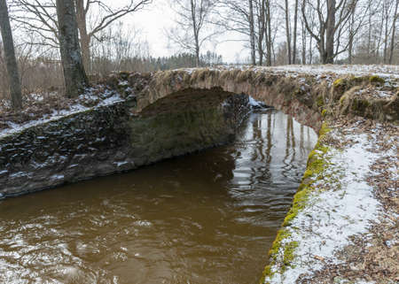 landscape with a continuous boulder stone bridge with a brick used for masonry, early spring, bare trees, snow plan on the ground, Stone arch bridge over the river Kuja, Madona, Latviaの写真素材