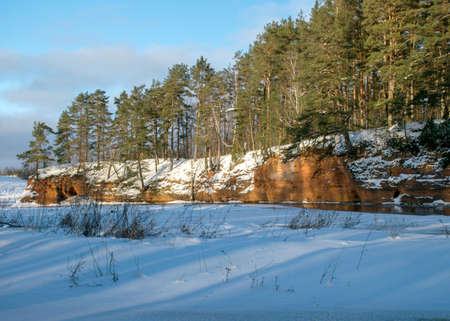 sunny winter landscape with red sandstone cliffs on the bank of the river Salaca, the sun shines on the trees and the river bank, the ice covers the river, Sarkanas clifts Latviaの写真素材