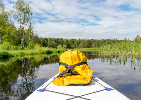 colorful summer landscape from the river, in the foreground a sup board and a yellow rubber bag, cloud reflections in the water, green trees and grass on the banks of the river, Sedas River, Latviaの写真素材