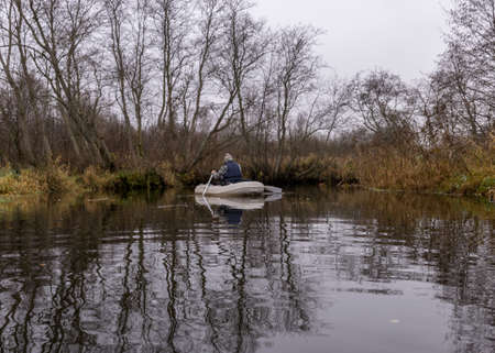 gray and cloudy day, fisherman in a white boat, river bank with bare trees and bushes, shore reflection in river water, autumnの写真素材