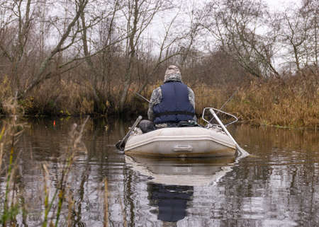 gray and cloudy day, fisherman in a white boat, river bank with bare trees and bushes, shore reflection in river water, autumnの写真素材