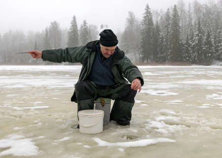 landscape with a man in winter who sits on the ice next to a hole and holds a small fishing rod. A fisherman is sitting on a frozen lake and fishing, winter dayの写真素材