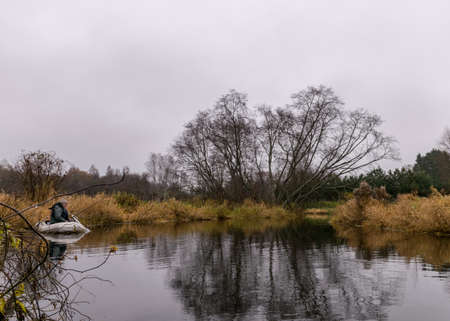 gray and cloudy day, fisherman in a white boat, river bank with bare trees and bushes, shore reflection in river water, autumnの写真素材