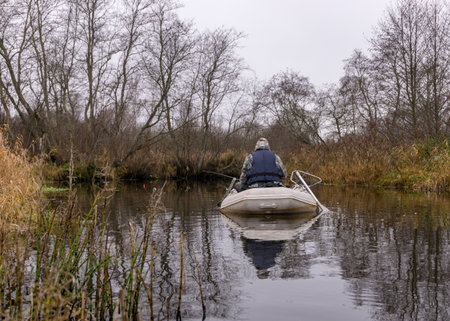 gray and cloudy day, fisherman in a white boat, river bank with bare trees and bushes, shore reflection in river water, autumnの写真素材