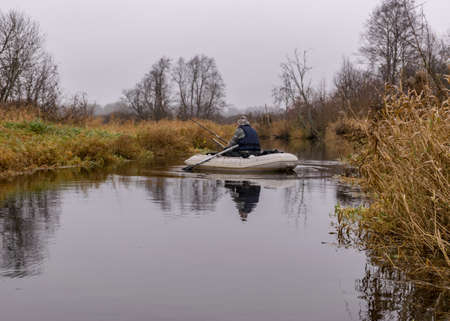 gray and cloudy day, fisherman in a white boat, river bank with bare trees and bushes, shore reflection in river water, autumnの写真素材