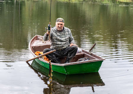 landscape with a fisherman in a boat, a small pike attached to the fisherman, a calm lake water surface, tree reflections, a cloudy day, fishing conceptの写真素材