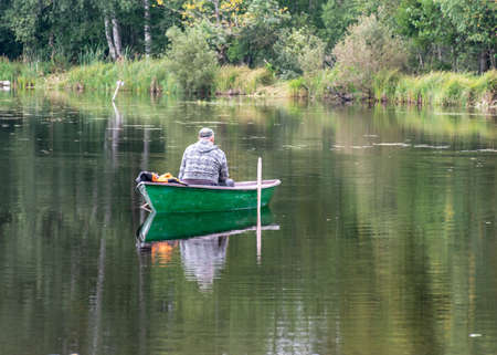 landscape with a fisherman in a boat, calm lake water surface, tree reflections, cloudy day, fishing conceptの写真素材