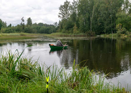 landscape with a fisherman in a boat, calm lake water surface, tree reflections, cloudy day, fishing conceptの写真素材