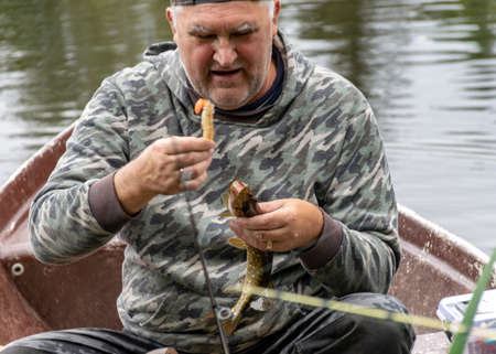 landscape with a fisherman in a boat, a small pike attached to the fisherman, a calm lake water surface, tree reflections, a cloudy day, fishing conceptの写真素材