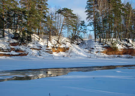 sunny winter landscape with red sandstone cliffs on the bank of the river Salaca, the sun shines on the trees and the river bank, the ice covers the river, Sarkanas clifts Latviaの写真素材