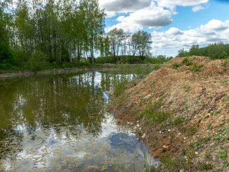 landscape with a fish pond, in the water of which there are colorful cumulus clouds and trees, there are recently dug gravel piles on the shore of the pond, the first spring greenery in natureの写真素材