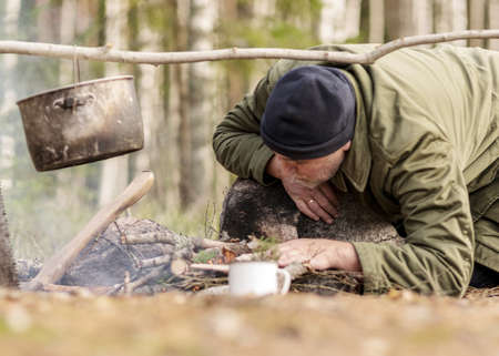 man in the forest trying to light a campfire, a tree branch with a pot over the campfire, blurred forest background, bonfire and smoke, autumn time in natureの写真素材