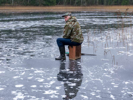 winter angler fishing on ice, reflections on the ice surface, naked tree silhouettes on the shore, ice texture in the foregroundの写真素材