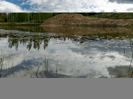 landscape with a fish pond, in the water of which there are colorful cumulus clouds and trees, there are recently dug gravel piles on the shore of the pond, the first spring greenery in natureの写真素材