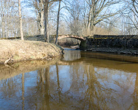 The Masonry bridge bridge over the river of Cuja, a sunny day in early spring with clear blue skies, the banks of a small river overgrown with last year's grass. tree reflections in water, Kuja river, Latviaの写真素材