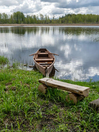 beautiful landscape with calm lake water, white cumulus clouds and reflections in the water, brown wooden boat on the lake shore, wooden bench in the foregroundの写真素材