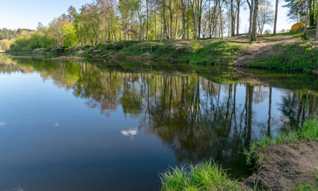 colorful spring landscape with a river, in the waters of which clouds are reflected, the banks of...