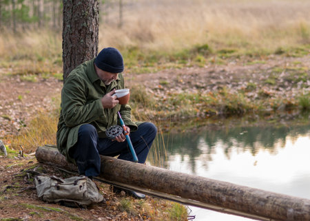 fisherman by the river sits on a tree branch and drinks tea, blurred tree background, autumn timeの写真素材