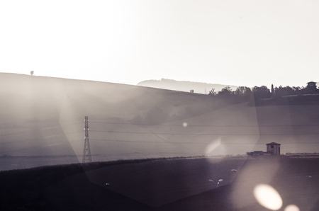 Sun appears over the hill to enlighten an electric tower and a small house in the middle of a fieldの写真素材