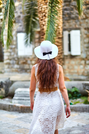 Young brown-haired woman in white hat and dress walking the streets of the old cityの写真素材
