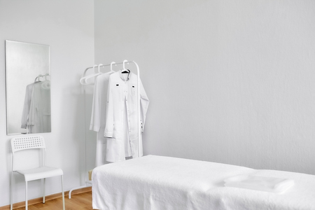 Consulting room. Cosmetic couch, chair, table, hanger with doctor's dressing gowns. Interior with white walls.の写真素材