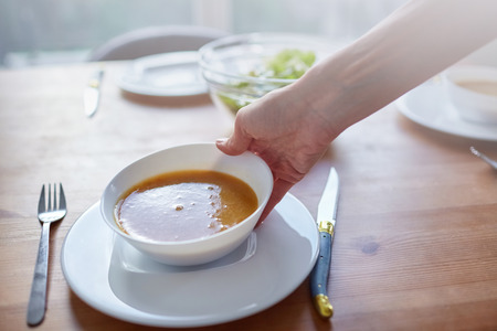 Female hand with a white bowl of vegetable soup. Serving dishes at the dining tableの写真素材