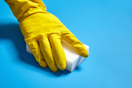 Human hand in yellow rubber glove holds a white melamine household sponge on blue backgroundの写真素材