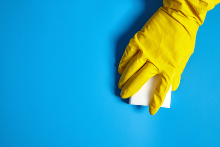 Human hand in yellow glove holds a white melamine sponge on blue background with copy space. Cleaning homeの写真素材