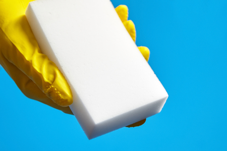 Human hand in yellow glove holds a white melamine sponge on blue background. Universal tool for cleaning various surfaces from dirt and stainsの写真素材