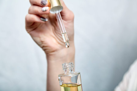 Pipette with drop of cosmetic oil and bottle in female hand, closeup