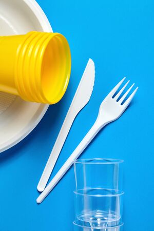 Disposable plastic tableware on blue background. White plate forks, knives, yellow and transparent plastic glassesの写真素材