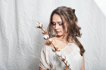Portrait of young woman with cotton plant with fluffy flowers on white background. Attractive lady with long wavy brown hairの写真素材