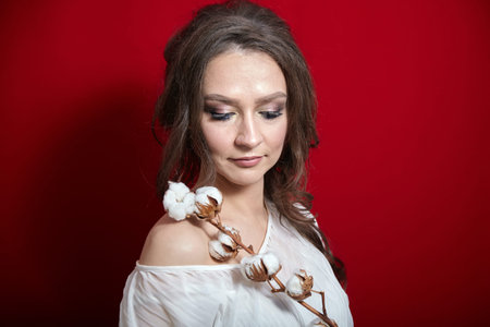 Portrait of young woman with cotton plant with white fluffy flowers on red background. Attractive lady with long wavy brown hairの写真素材