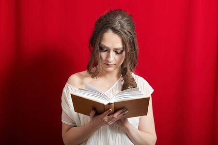 Young woman reading book in leather cover on the background of red curtainsの写真素材