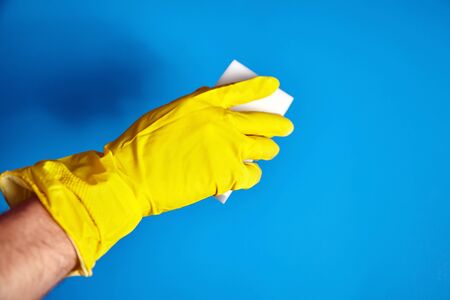 Hand in yellow protective glove with white melamine household sponge on blue background. Means for cleaning surfaces from dirt and stainsの写真素材