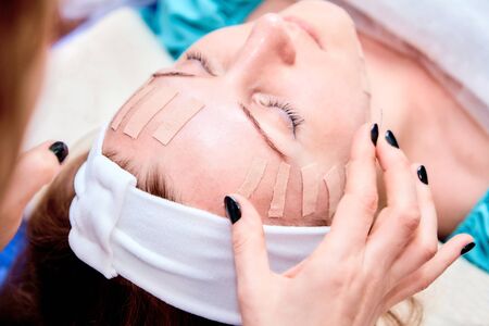 Face aesthetic taping. Facial skin care. Young woman lying with tape applications on face. Non-invasive anti-ageing lifting method for reduction of wrinkles on the forehead and the outer eye cornersの写真素材