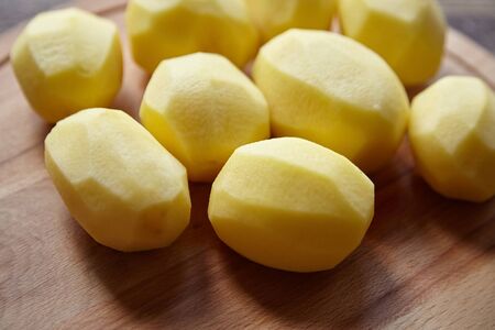 Peeled potatoes on wooden cutting board on brown table. Cooking food from natural products. Root vegetable. Raw ingredient: uncooked whole potatoesの写真素材