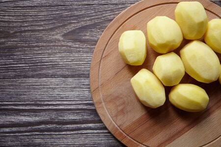 Peeled potatoes on wooden cutting board on brown table. Cooking food from natural products. Root vegetable. Raw ingredient: uncooked whole potatoesの写真素材