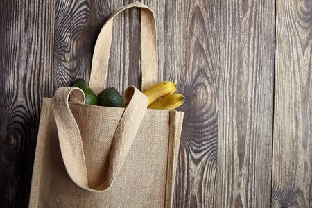 Reusable shopping bag with fresh fruits. Natural eco friendly material. Yellow bananas, green avocado, hessian or jute sack on brown wooden table. Useful organic productsの写真素材