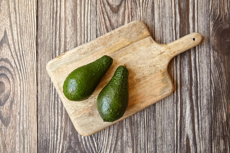 Avocados on a natural pine cutting board on wooden background. Two whole green fresh tropical fruits...