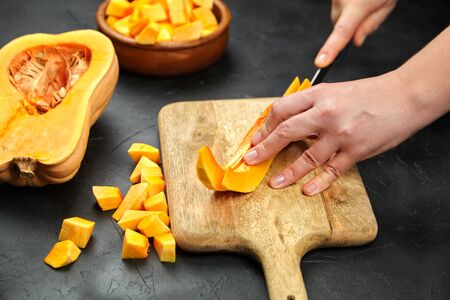 Woman cuts a pumpkin on wooden chopping board on stone table. Female hands with steel knife, butternut squash pieces in wooden bowl on black background. Cooking sweet pumkinの写真素材