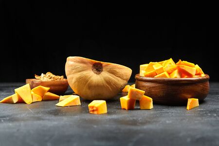 Pumpkin half, fresh pieces in wooden bowl and scattered on black background. Chopped raw butternut squash, selective focus. Cooking dish from vegetable ingredients, plant-based foodの写真素材